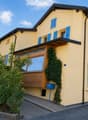 Two-story yellow house with blue shutters, wooden front door, and ivy-covered wall. The house features a balcony and is surrounded by greenery and a parked car.