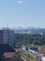 A panoramic view of a cityscape with a mix of modern and traditional buildings, surrounded by green fields and distant mountains under a clear blue sky.