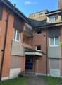 Multi-story brick apartment building with multiple windows, balconies, and a small garden area in front.
