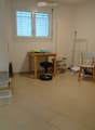 A small, furnished room with a tiled floor, a wooden table with a chair and stool, a bookshelf filled with books and binders, and several storage bins along the walls.