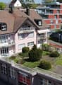Aerial view of a two-story residential building with a pink facade, multiple balconies, and a well-maintained garden area. The building features a garage and parking area, with neighboring structures visible in the background.