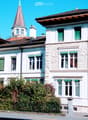 A large, white stone building with multiple stories, green shutters, and a prominent tower with a pointed roof. The building features a balcony and is surrounded by greenery and a fence.