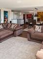 Modern living room with large windows offering a view of greenery, featuring a brown sectional sofa, ottoman, dining area with table and chairs, and decorative art pieces.