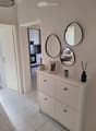 Hallway with white tiled floor, light-colored walls, and a small console table with decorative items, mirrors, and a view into a kitchen and bedroom area.