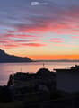 View from a balcony or terrace overlooking a lake and mountains at sunset, with city buildings in the foreground.