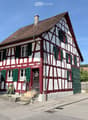 Traditional half-timbered house with red and white exterior, green shutters, and a small garden area with potted plants near the entrance.