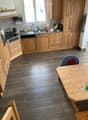 The kitchen features light wooden cabinets, a granite countertop, and a wooden dining table with a single chair. The floor is made of wood, and the sink area includes a faucet and various small items.
