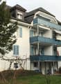 A multi-story residential building with light blue shutters, balconies, and a well-maintained lawn.