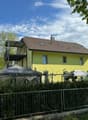 Two-story yellow house with a brown tiled roof, multiple windows, and a balcony on the upper level. The house is surrounded by greenery and a metal fence.