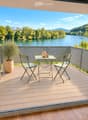 Balcony with wooden decking, metal railing, table with two chairs, vase, and cup, overlooking a river and green landscape.