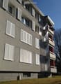 Four-story residential building with grey facade, white shutters, and multiple balconies with green awnings.