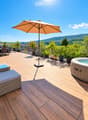 Wooden deck with a sofa, glass coffee table, dining table with chairs, umbrella, and a hot tub, surrounded by potted plants and offering a scenic view of the valley.