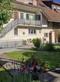 Well-maintained garden with a wooden deck, potted plants, and a metal sculpture. The house features a balcony, multiple windows, and a staircase leading to the entrance.