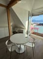 Bright attic room with wooden floors, white furniture, and a large window offering a scenic view of the lake and mountains. The room features a round white table with two chairs, a desk with a chair, and a white cabinet.