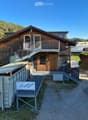 Two-story wooden house with a balcony, staircase, and a small shed with a ladder. The house is surrounded by greenery and has a clear view of mountains in the background.