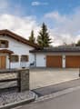 A two-story house with a white exterior, wooden garage doors, and a stone driveway. The house features a traditional alpine design with wooden shutters and a sloped roof. There is a decorative wooden fence and a small statue near the entrance.