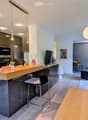 Modern open-plan living and dining area with a kitchen island, bar stools, sofa, coffee table, and dining table. The kitchen features dark cabinets, a sink, and an oven. The living area includes a sofa, a coffee table, and a TV screen.