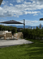 A modern outdoor patio with a wooden deck, white sofa, two wicker chairs, and a large umbrella, surrounded by greenery and a scenic mountain view.