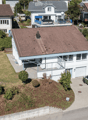 Aerial view of a modern two-story house with a tiled roof, white exterior, and a garage. The house features a raised ground floor, a balcony, and is surrounded by greenery and neighboring houses.