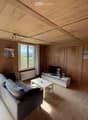 Bright living room with wooden walls and ceiling, large windows offering a scenic view, grey sofa, white coffee table, TV stand, and a small flower vase.