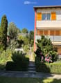 Two-story house with yellow shutters, balcony, garden, and steps leading to the entrance.