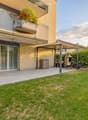 Well-maintained garden with a neatly trimmed lawn, a patio area with a pergola, a barbecue, and a small storage shed. The house features a balcony with plants and flowers, and the exterior is made of light-colored brick.