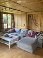 Cozy living room with light wooden walls and ceiling, grey sectional sofa with purple and grey cushions, white coffee table with remote controls and a basket, floor lamp, and decorative picture frames on the wall.