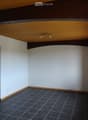Empty room with white walls, dark tiled floor, wooden ceiling beams, and a window with blinds.