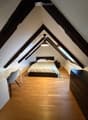 Cozy attic bedroom with wooden floors, slanted ceilings, and large windows. Features a bed with white bedding, a nightstand, a desk with a chair, and a hanging pendant light.