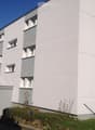 Modern multi-story building with white facade, grey accents, and multiple windows with shutters. Small garden area in front with some plants and a clothesline.