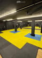 A spacious gym area with a large yellow and blue mat, boxing bags hanging from the ceiling, weights, a bench, and a mirror on the wall.
