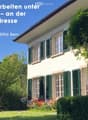 A two-story house with white walls, green shutters, and a well-maintained garden with red flowers and green shrubs.