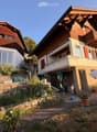 Two-story house with wooden exterior, red shutters, and a balcony. The garden features potted plants, a stone wall, and a patio area with outdoor furniture.