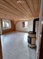 Empty attic room with wooden slanted ceiling, tiled floor, and a fireplace in the corner. Two windows provide natural light.