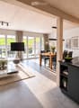 Open-plan living and dining area with a modern sofa, coffee table, dining table, and kitchen island. Large windows and sliding doors lead to an outdoor patio.