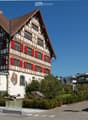 Traditional Swiss-style building with a steeply pitched roof, exposed wooden beams, and red shutters. The structure is two stories with multiple windows and a small balcony on the upper level.