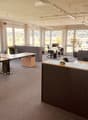 Bright office space with large windows, multiple desks, chairs, and office equipment.