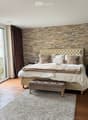 Elegant bedroom with a large bed, fireplace, mirror, and decorative lamp. Features a stone accent wall and large window with curtains.