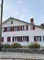 Two-story white house with red shutters, multiple windows, chimney, and a small garden behind a metal fence.