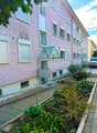 Pink residential building with multiple windows, a small garden area with plants and flowers, and a parked car in the background.