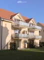 Two-story residential building with beige walls, red-tiled roof, multiple windows, and balconies with glass railings.