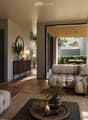 Modern living room with a light grey sectional sofa, coffee table, floor lamp, side table, mirror, bookshelf, and potted plants. The space features large windows and a view of an outdoor area.