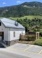 Single-story house with a grey metal roof, white walls, and a small staircase leading to the entrance. The house is situated in a scenic valley with vineyards and mountains in the background.