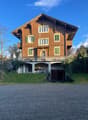 A two-story wooden house with green shutters, a balcony, and a small garage. The house features a pitched roof and is surrounded by a well-maintained garden.