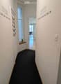 A clean, well-lit hallway with a dark carpeted floor, white walls, and a small window with blinds. There is a coat rack on the left and a small plant on a shelf near the window. The hallway leads to an open area with more rooms visible in the background.