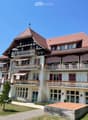 A large, multi-story house with a red-tiled roof, white walls, and multiple balconies adorned with striped awnings and flower boxes. The house features a mix of traditional and modern architectural elements, including wooden beams and large windows.