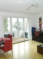 Bright living and dining area with wooden flooring, large windows, and a balcony access. Features a dining table with red chairs, a black cabinet, a sofa, and decorative items on the windowsill.
