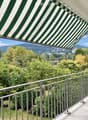 Balcony with metal railing, green and white striped awning, overlooking a lush green landscape with trees and distant hills.