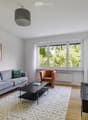 Modern living room with a grey sofa, orange armchair, black coffee table, TV stand, and a large window with white curtains.