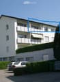 A modern, multi-story residential building with white exterior walls, multiple balconies, and a well-maintained garden area. The building features large windows and a clean, contemporary design.